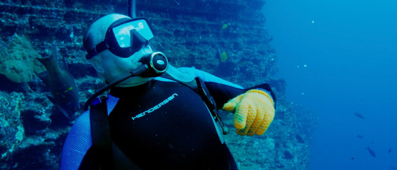 Oudere duiker met gele handschoenen en volledig duikpak wijst naar iets naast een onderwaterrifwand