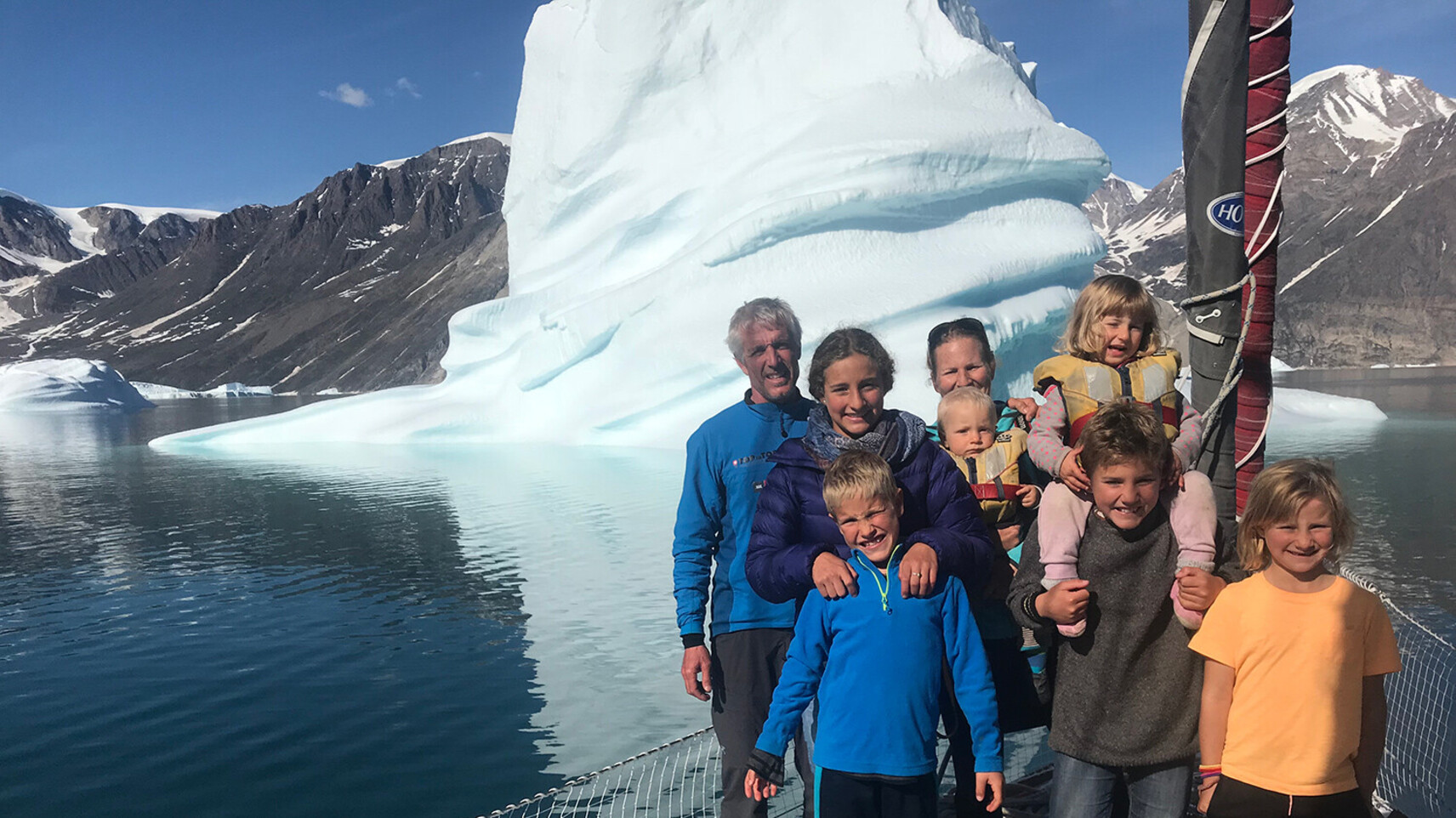 La famille Schw&ouml;rer pose ensemble sur le pont de leur voilier d'exp&eacute;dition TOPtoTOP devant un immense iceberg dans un fjord groenlandais &ndash; parents et six enfants lors d'un voyage de sensibilisation climatique